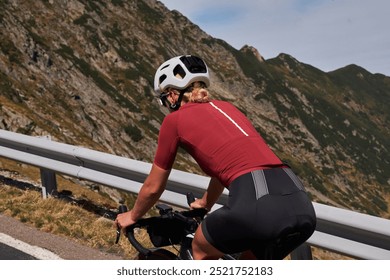 Female cyclist in a red jersey and black shorts rides a bike up a mountain road, surrounded by scenic mountains. Cyclist is wearing a helmet and is focused on the road ahead. Cycling hobby. - Powered by Shutterstock