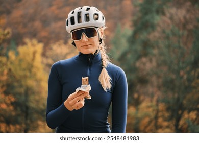 Female cyclist takes a break to enjoy a healthy energy bar. Autumnal forest background. Active lifestyle, fitness, and healthy eating concept. Sport snack for cyclist. Granola energy bar. - Powered by Shutterstock