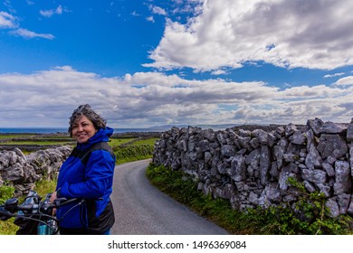 Female cyclist taking a break on a country road, Irish fields with stone fences with the sea in the background on Inis Oirr Island, gray short hair, blue jacket, sunny day in Aran Islands, Ireland - Powered by Shutterstock