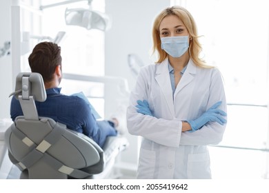 Female Dentist Blonde Woman In Protective Face Mask And Medical Gloves Posing At Modern Dental Clinic, Unrecognizable Man Patient Sitting In Dental Chair, Dental Care While Coronavirus Epidemic