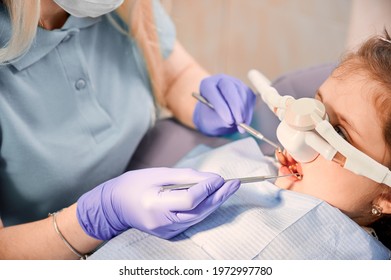 Female dentist checking child teeth with dental explorer and mirror while girl lying in chair with inhalation sedation at dental office. Concept of pediatric, sedation dentistry and dental care. - Shutterstock ID 1972997780