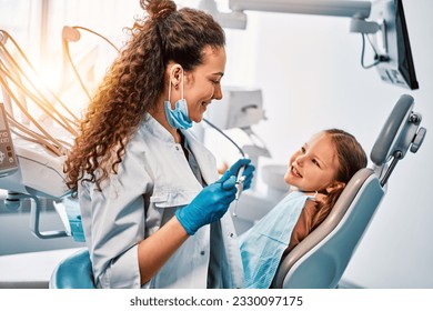 A female dentist doctor talks to a small patient, the child shows her teeth and sits in the dental chair.Sunlight. - Powered by Shutterstock