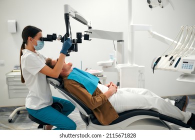 A Female Dentist Examines A Young Patient Looking At His Teeth With A Professional Microscope In A Dental Surgery Room. Dental Operating Microscope. Dental Optics.