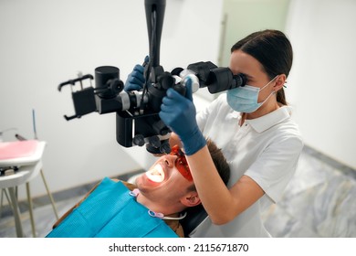 A Female Dentist Examines A Young Patient Looking At His Teeth With A Professional Microscope In A Dental Surgery Room. Dental Operating Microscope. Dental Optics.