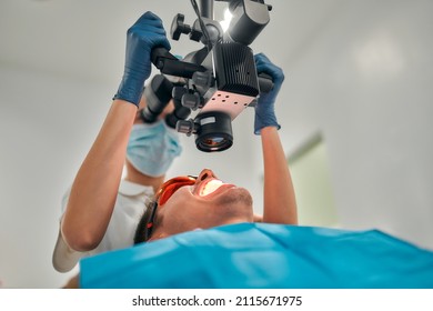 A Female Dentist Examines A Young Patient Looking At His Teeth With A Professional Microscope In A Dental Surgery Room. Dental Operating Microscope. Dental Optics.