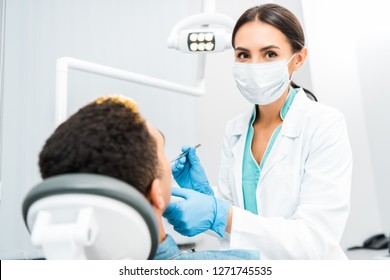 Female Dentist In Latex Gloves And Mask Examining African American Patient 