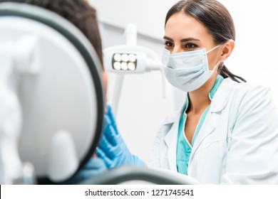 Female Dentist In Mask Looking At Patient In Dental Clinic