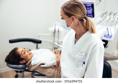 Female Dentist Wearing Face Mask Talking With Patient In Dentist's Chair. Dental Doctor With Female Patient Visiting For Routine Checkup.