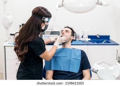 Female Dentist Wearing Face Shield Isolation Mask Working With Male Patient And Putting Teeth Covers.