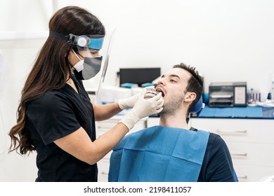 Female Dentist Wearing Face Shield Isolation Mask Working With Male Patient And Putting Teeth Covers.