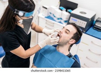 Female Dentist Wearing Face Shield Isolation Mask Working With Male Patient And Putting Teeth Covers.