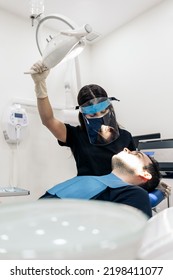 Female Dentist Wearing Face Shield Isolation Mask Using Light And Working In Modern Clinic.