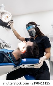 Female Dentist Wearing Face Shield Isolation Mask Using Light And Working In Modern Clinic.