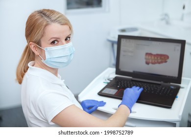 Female Dentist Wearing Medical Face Mask, Looking To The Camera While Working On Her Computer