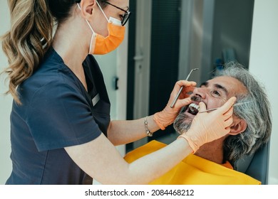 Female Dentist Working In Dental Clinic With Male Patient In The Dentist's Chair. 
Woman Dentist With Orange Protective Face Mask And Gloves Examining Her Mature Patient Teeth In Dentist's Office.