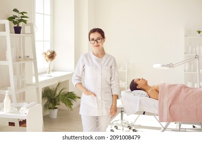 Female Dermatologist, Skin Therapist, Beautician And Skincare Professional With Patient In Office. Beautiful Woman In Coat Uniform And Glasses Standing In Room For Examinations And Cleaning Procedures