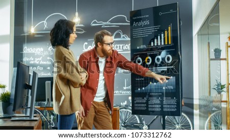 Female Developer and Male Statistician Use Interactive Whiteboard Presentation Screen to Look at Charts, Graphs and Growth Statistics. They Work in the Stylish Creative Office.