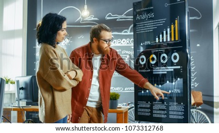 Female Developer and Male Statistician Use Interactive Whiteboard Presentation Touchscreen to Look at Charts, Graphs and Growth Statistics. They Work in the Stylish Creative Office.