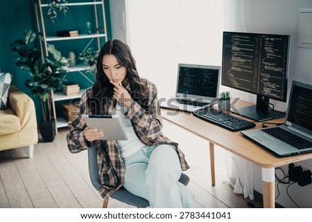 Female developer working remotely in a modern home office, concentrating on coding projects across multiple monitors and a tablet. Casual, comfortable workspace with plants and shelving conveying productivity and focus.