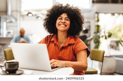 Female digital nomad smiling happily as she types on her laptop at a coffee house. Afro haired woman dressed in casual clothing and working online in a relaxed environment. - Powered by Shutterstock