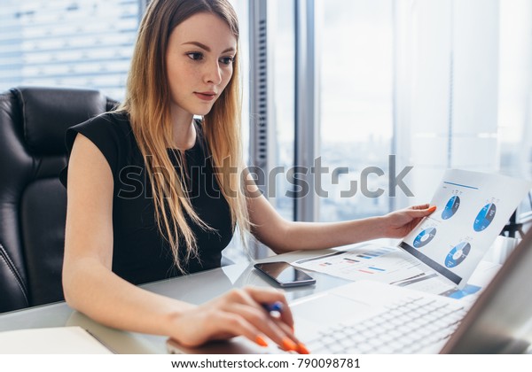 Female director working in office sitting at desk
analyzing business statistics holding diagrams and charts using
laptop