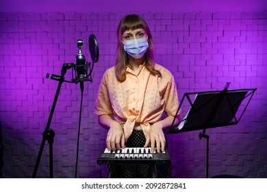 A Female DJ Stands On Stage Behind A Keyboard, A Music Stand And A Microphom In A Protective Mask. Party During The Pandemic