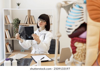 Female doctor analyzing X-ray in modern medical office. Medical professional wearing white coat and glasses, working on patient diagnosis. Room features anatomical model, bookshelves, and desk. - Powered by Shutterstock