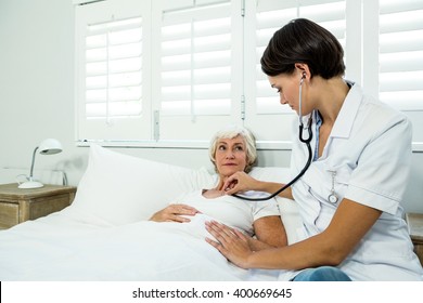 Female Doctor Auscultating Aged Woman Against Window At Home