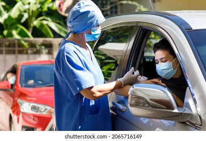 Female Doctor In Blue Hospital Uniform And Face Mask Stand Near Car Holding Syringe In Hands Wears Rubber Gloves Injecting Covid 19 Vaccine To Drive Through Patient Shoulder On Vaccination Queue.