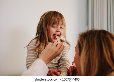 Female Doctor Checking For Sore Throat Of Girl Patient. Girl Child Having Throat Examination With Tongue Depressor By A Pediatric Doctor.