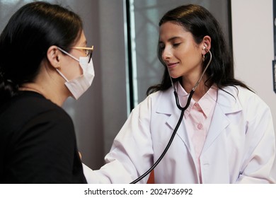 Female Doctor Checks Patients Heart Beat And Lungs Using Stethoscope. Woman Doing General Health Check Up Wearing Disposable Face Mask