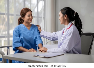 A female doctor is comforting a female patient in a hospital setting. The doctor is wearing a white lab coat and the patient is wearing a blue scrubs. The doctor is holding the patient's hand - Powered by Shutterstock