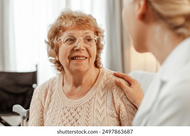 Female doctor comforting a smiling senior woman recovering from injury at home. They are both wearing casual clothes and the woman is looking away. Treatment, health care concept - Powered by Shutterstock