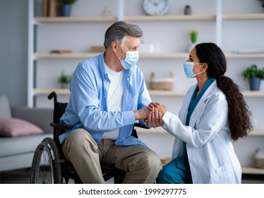 Female Doctor And Disabled Elderly Man In Wheelchair Wearing Protective Masks During Covid Lockdown, Holding Hands At Retirement Home. Caregiver And Impaired Patient Staying Safe Together