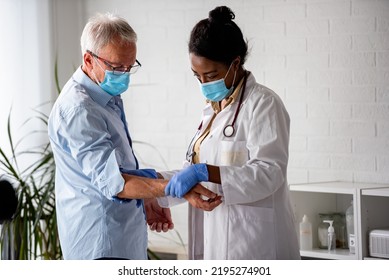 Female Doctor Is Examining Male Elderly Patient At Clinic. Doctor And Patient Wearing A Face Mask.