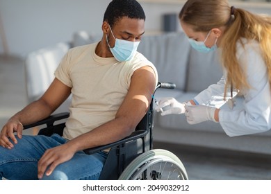 Female Doctor Giving Covid-19 Vaccine Or Medication Shot To Paraplegic Black Male Patient In Wheelchair At Home. African American Guy In Face Mask Receiving Coronavirus Immunization Indoors