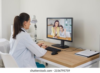 Female Doctor Giving Telemedicine Consultation To Happy Family Via Videocall. Young Paediatrician Sitting At Office Desk And Using Desktop Computer To Have Online Conversation With Mom And Her Child