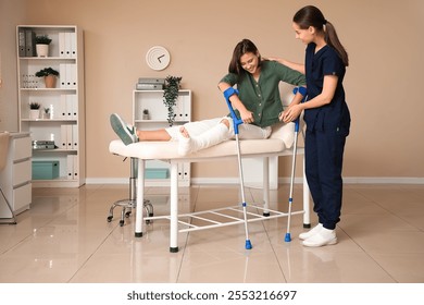 Female doctor helping woman with broken leg and crutches to stand up from couch in clinic - Powered by Shutterstock