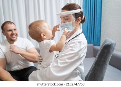 Female Doctor Holding Baby, Reaching For Her Face Shield