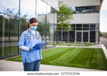 Female doctor holding blue clipboard standing outside hospital or clinic,frontline key medical worker filling out patient card form,exterior entrance to a modern care facility or nursing home complex