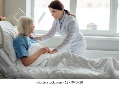 Female Doctor Holding Hands And Supporting Sad Senior Woman Lying In Hospital Bed