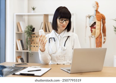 Female doctor holding pill bottle while using laptop at desk. Professional healthcare worker consulting online or researching medication. Bright and modern medical setting with anatomical model. - Powered by Shutterstock