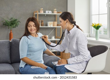 Female doctor listens to the heart and breath of a young woman through a stethoscope on her chest. Woman sitting on sofa at home. Concept of medical care and calling the doctor home.