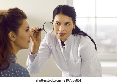 Female Doctor Looking At Patient's Skin Through Magnifying Glass. Professional Dermatologist And Skincare Specialist Investigating Moles Or Tumor Growth Signs On Woman's Face. Skin Checkup Concept