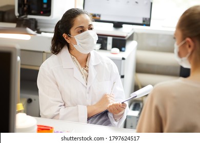 Female Doctor In Mask Making Notes In Medical Card While Talking To Patient At Hospital