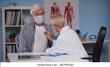 Female Doctor In Mask Using Stethoscope To Examine Heart Of Senior Man Patient. Aged Male Patient Visiting Practitioner Wearing Protective Mask And Being Examined With Stethoscope