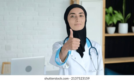 A Female Doctor In A Medical Gown And A Black Hijab Shows A Thumbs Up, A Muslim Woman At Her Workplace In An Office In A Hospital, A Muslim Woman Works As A Nurse, Helps People.