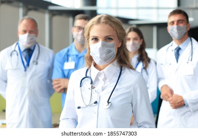 Female Doctor In Medical Mask With Group Of Happy Successful Colleagues Also In Preventive Masks.