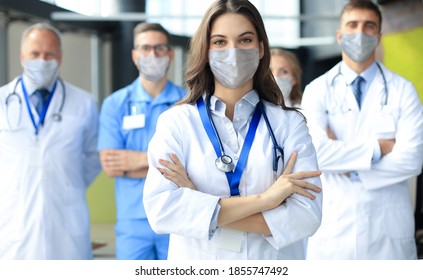 Female Doctor In Medical Mask With Group Of Happy Successful Colleagues Also In Preventive Masks.