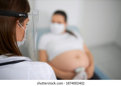 Female Doctor In Medical Protective Mask Doing Checkup With Ultrasound For Pregnant Woman. Concept Of Pregnancy And Pandemic Time.